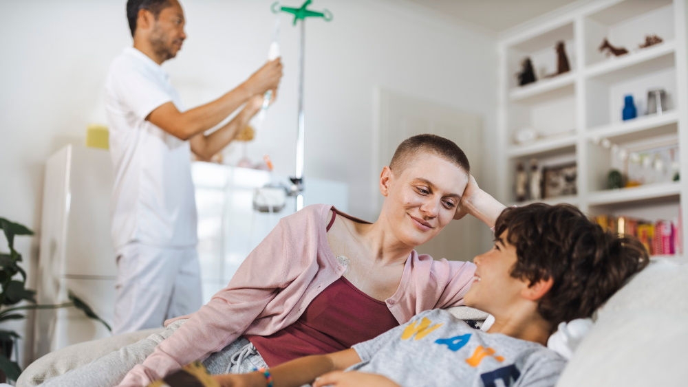 Eine Frau mit sehr kurzem Haar liegt neben einem Jungen auf einem Sofa, beide mit angehobenem Oberkörper. Sie schauen sich mit einem Lächeln an. Im Hintergrund hängt eine Krankenschwester einen Infusionsbeutel an ein Gestell.