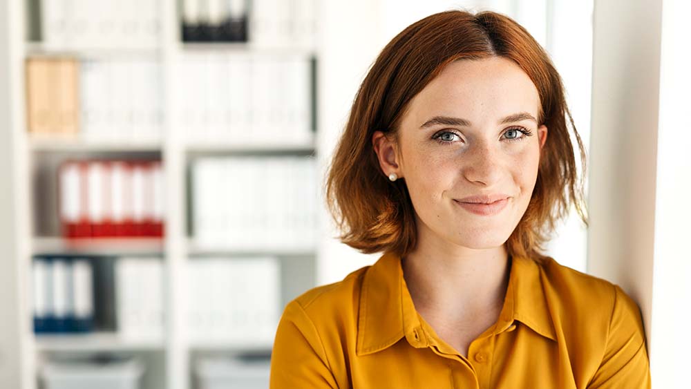 Eine Frau lächelt selbstbewusst, blickt direkt in die Kamera, im Hintergrund sind Bücherregale sichtbar. 